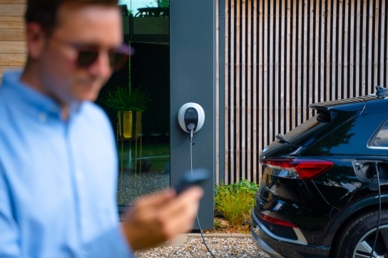 Man checking his mobile phone with a pod point charger in the back ground charging a black electric car